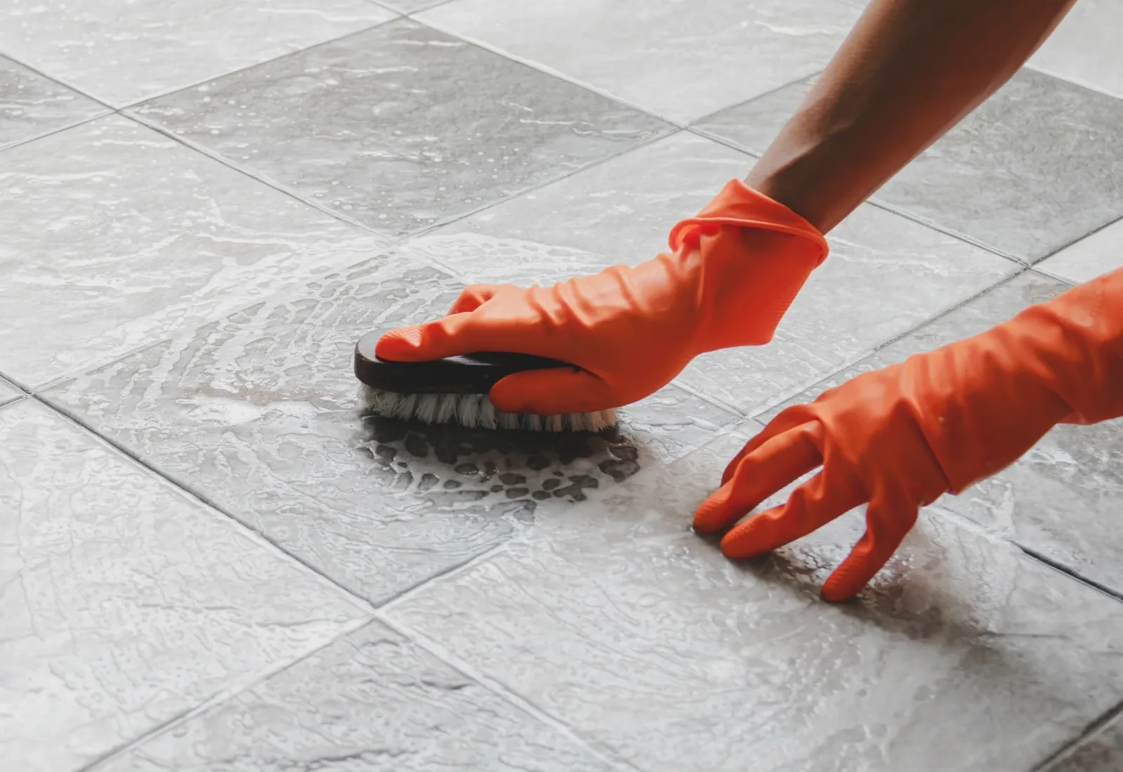 Gloved hand scrubbing grey floor tiles and grout with a brush